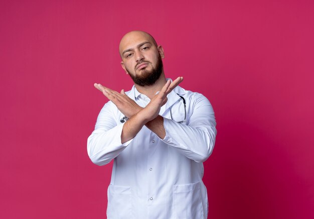 Strikte jonge kale mannelijke arts die medische mantel en stethoscoop draagt die gebaar van nee toont dat op roze wordt geïsoleerd