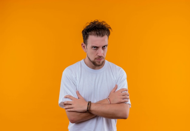 Strikte jonge kerel die een wit t-shirt draagt, gekruiste handen op geïsoleerde oranje achtergrond