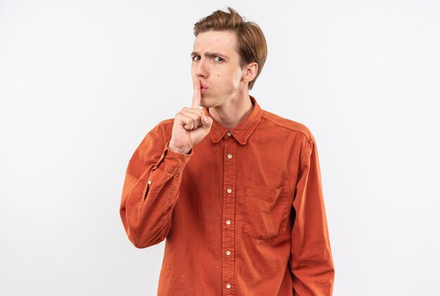 Strikte jonge knappe kerel die een rood overhemd draagt dat stiltegebaar toont dat op witte muur wordt geïsoleerd