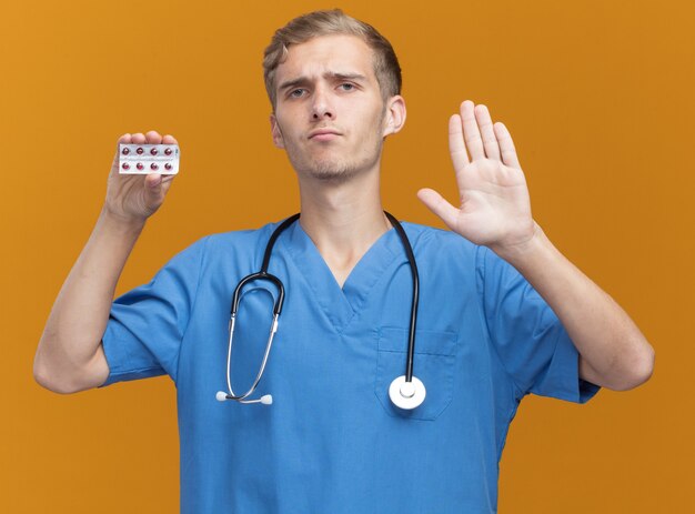 Strikte jonge mannelijke arts die arts eenvormig met de pillen van de stethoscoopholding draagt die eindegebaar tonen dat op oranje muur wordt geïsoleerd