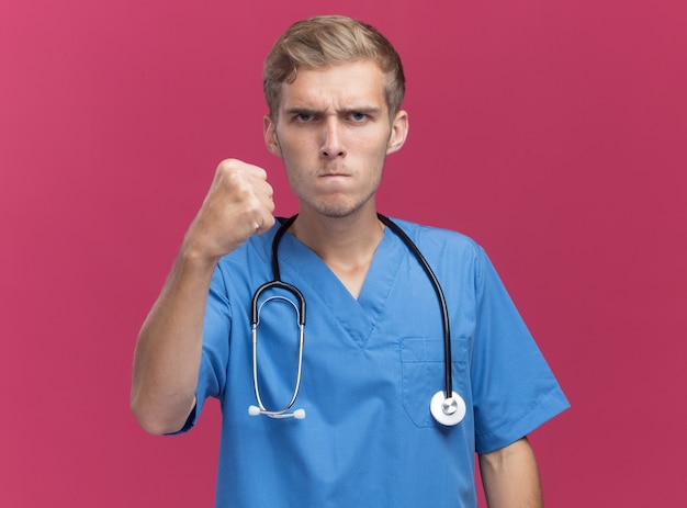 Strikte jonge mannelijke arts die artsenuniform met de vuist van de stethoscoopholding draagt die op roze muur met exemplaarruimte wordt geïsoleerd