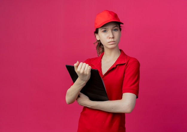 Strikte jonge mooie levering meisje in rood uniform en pet houden klembord kijken naar camera geïsoleerd op karmozijnrode achtergrond met kopie ruimte