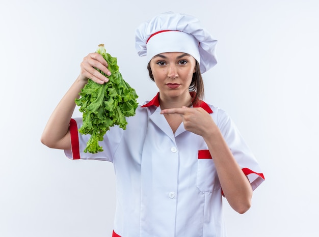 Strikte jonge vrouwelijke kok die een uniforme chef-kok draagt en wijst op salade die op een witte achtergrond wordt geïsoleerd