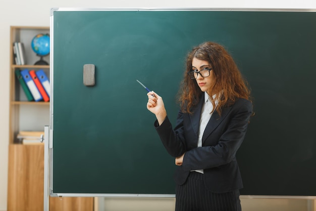 strikte jonge vrouwelijke leraar die vooraan staat, wijst naar schoolbord met aanwijzer in de klas