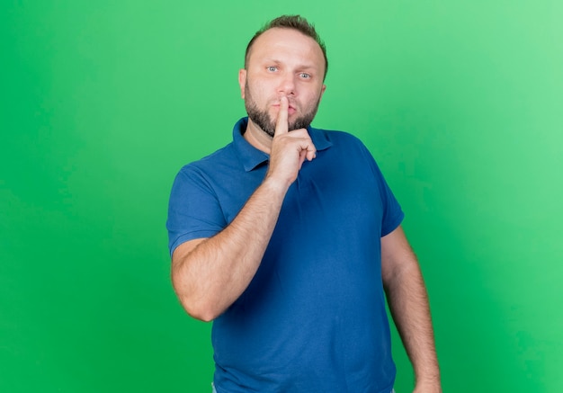 Strikte volwassen Slavische man en doet stilte gebaar geïsoleerd op groene muur met kopie ruimte