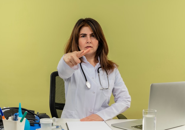 Strikte vrouwelijke arts van middelbare leeftijd die medische mantel draagt met een stethoscoop achter Bureau werkt op laptop met medische hulpmiddelen die je gebaar met kopie ruimte tonen