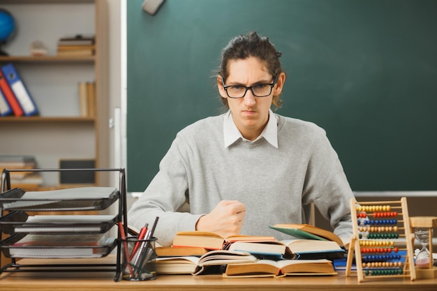 strikte vuist jonge mannelijke leraar zittend aan een bureau met schoolhulpmiddelen in de klas