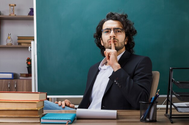 Strikte weergave van een mannelijke leraar met een stiltegebaar die een bril draagt die aan tafel zit met schoolhulpmiddelen in de klas