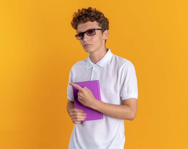 Gratis foto student man in casual kleding met een bril die een boek vasthoudt en naar de camera kijkt met een serieus gezicht wijzend met de vinger naar de zijkant die over een oranje achtergrond staat