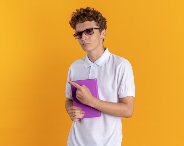 Student man in casual kleding met een bril die een boek vasthoudt en naar de camera kijkt met een serieus gezicht wijzend met de vinger naar de zijkant die over een oranje achtergrond staat