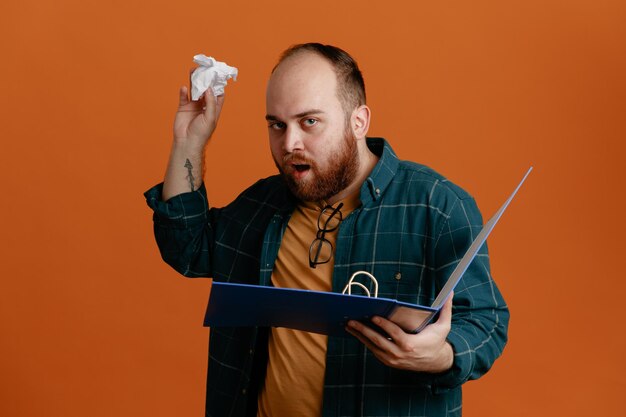 Student man in vrijetijdskleding met kantoormap verfrommeld papier en kijkt verbaasd over oranje achtergrond