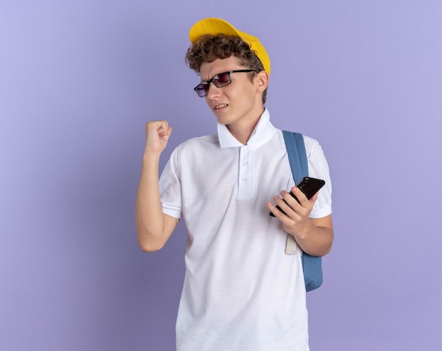 Student man in wit poloshirt en gele pet met bril met rugzak met smartphone balde vuist blij en opgewonden blij met zijn succes staande over blauwe achtergrond