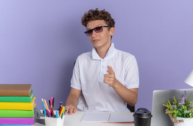 Student man in wit poloshirt met bril zittend aan de tafel met boeken die er zelfverzekerd uitziet wijzend met wijsvinger naar camera over blauwe achtergrond