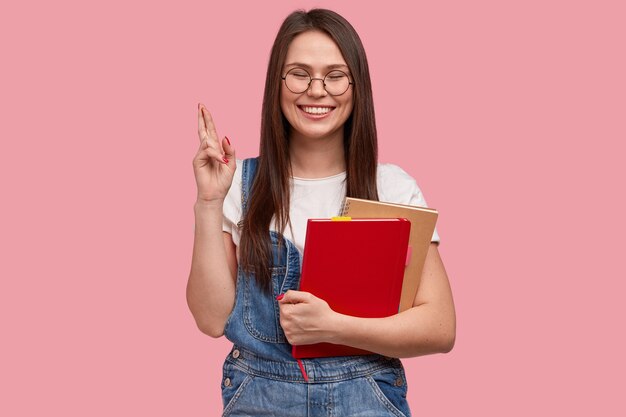 Student met vrolijke blik, kruist vingers voor geluk op examen, houdt notitieboekje voor het schrijven van records, gekleed in denim overall