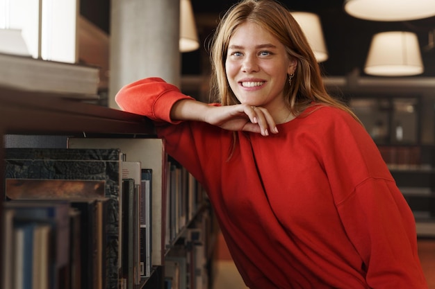 Studenten carrière en universiteit concept Dromerige mooie jonge vrouw met rood haar leunt op boekenplank glimlachend wegkijkend naar raam in bibliotheek studeren droom om een A-test of examen te krijgen