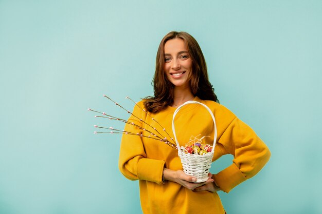 Studio die van een jonge Europese vrouw met overhemdskapsel is ontsproten die lichte kleren dragen die met de mand van vakantiePasen stellen over blauwe muur stellen