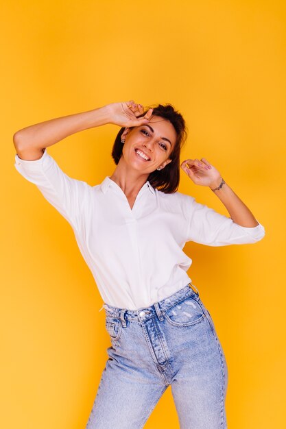 Studio die van gelukkig vrouwen kort haar is ontsproten die wit overhemd en denimbroek dragen die op gele muur stellen