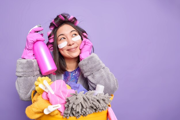 Studio-opname van een Aziatische vrouw met een dromerige uitdrukking past collageenpleisters toe onder de ogen houdt wasmiddel gekleed in huishoudelijke kleding poses in de buurt van een mand vol wasgoed geïsoleerd over paarse muurkopieruimte