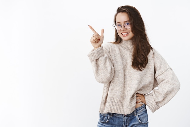 Studio-opname van een charmante, slimme en zelfverzekerde vrouw die een keuze maakt, wijzend naar de linkerbovenhoek om aan te geven dat het product breed naar voren lacht, wetende dat ze opgetogen over een grijze muur wil staan.