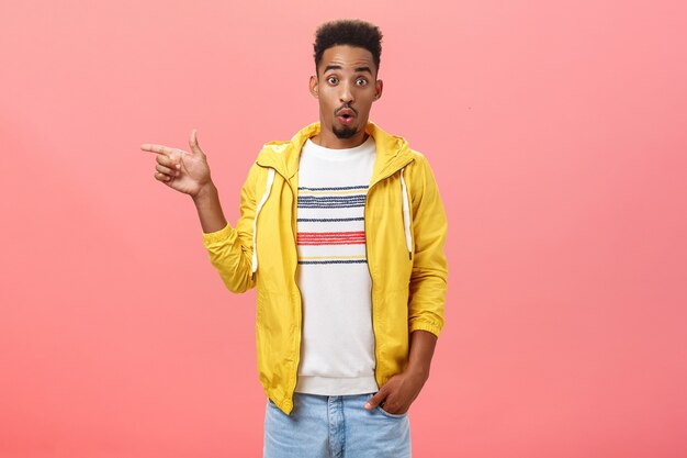 Studio-opname van verraste onder de indruk stijlvolle Afro-Amerikaanse mannelijke student in trendy gele jas die lippen vouwt in wow-geluid dat wenkbrauwen opheft van verbazing en naar links wijst, ondervraagd over roze muur