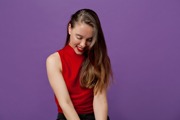 Studio portret van aantrekkelijke mooie vrouw met bruin haar, gekleed in rode top naar beneden te kijken en glimlachend op violet