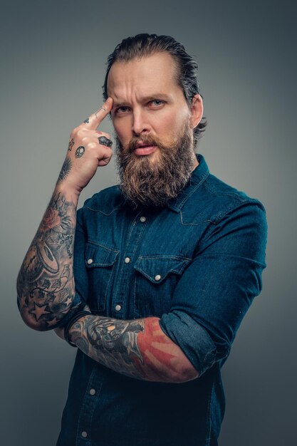 Studio portret van bebaarde man met tatoeages op zijn armen, gekleed in een denim shirt geïsoleerd op grijze vignet achtergrond.