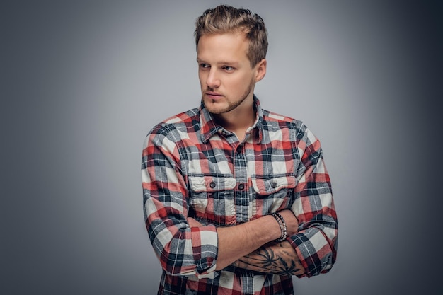 Studio portret van blonde man met gekruiste getatoeëerde armen, gekleed in een geruite overhemd geïsoleerd op grijze vignet achtergrond.