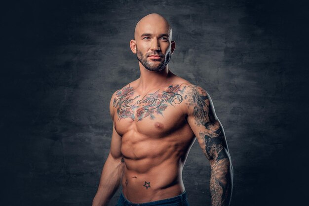 Studio portret van shirtless atletische geschoren hoofd man met tatoeages op zijn torso poseren over grijze vignet achtergrond.