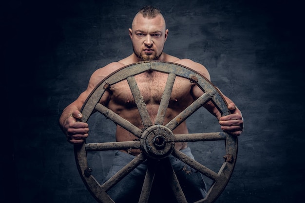 Studio portret van shirtless gespierde slaaf man houdt houten wiel over grijze achtergrond.