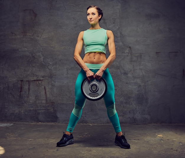 Gratis foto studio portret van sportieve vrouw gekleed in een azuurblauwe sportkleding uit te werken met barbell gewicht over grijze achtergrond.