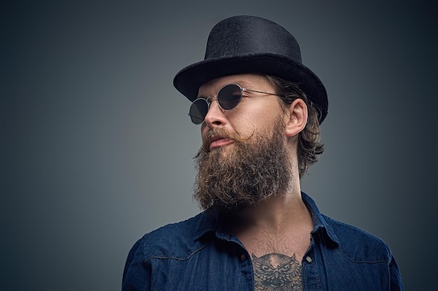 Studio portret van stijlvolle bebaarde man met een tatoeage op zijn borst, gekleed in een denim shirt, cilinder hoed en zonnebril geïsoleerd op een grijze achtergrond.