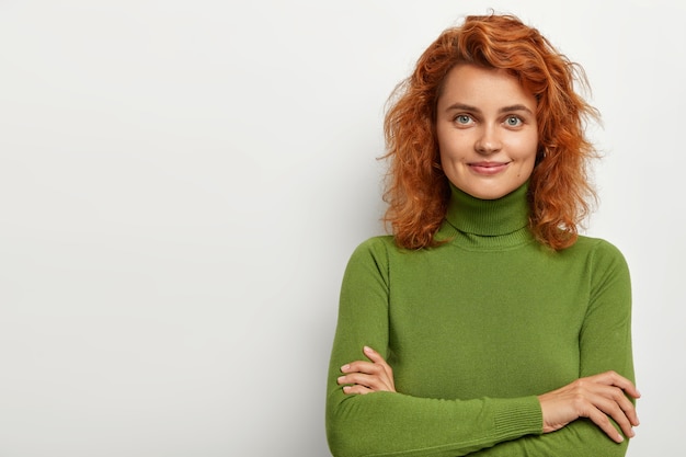 Studio shot van aantrekkelijk jong vrouwelijk model met kort rood krullend haar, gezonde huid, heeft zachte glimlach