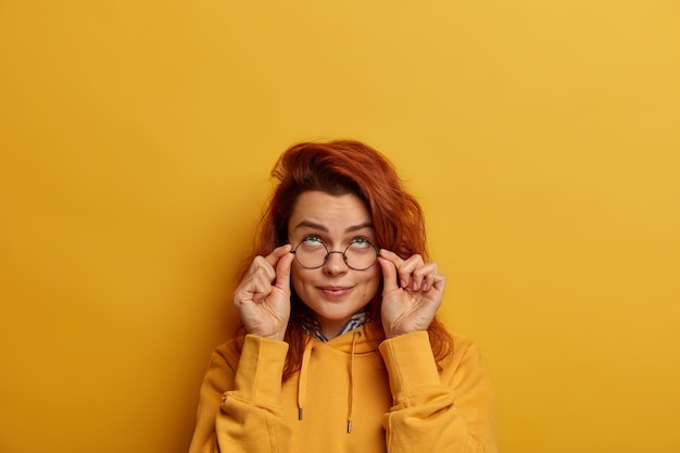 Studio shot van attente mooie europese vrouw kijkt serieus door ronde glazen, hierboven geconcentreerd