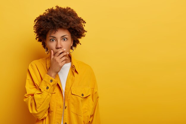 Studio shot van bang gekrulde vrouw bedekt mond met schrik, bang door angstaanjagend ding, gekleed in modieuze gele jas, kan ogen niet geloven, staat binnen, lege kopie ruimte voor advertentie