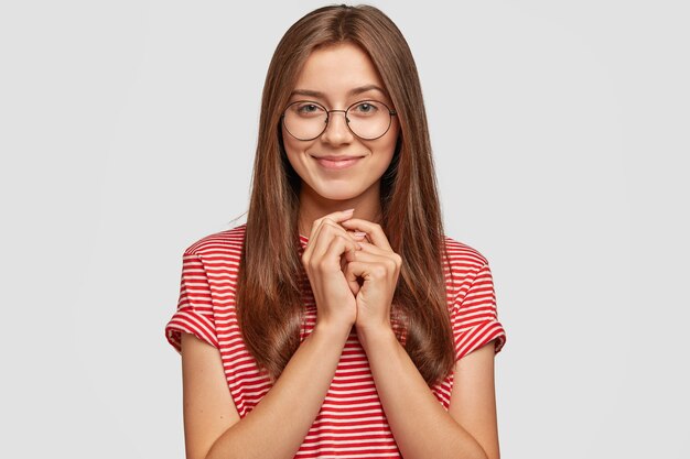 Studio shot van blanke vrouw met donker haar, aantrekkelijk uiterlijk, houdt de handen bij elkaar, kijkt met mysterieuze uitdrukking,