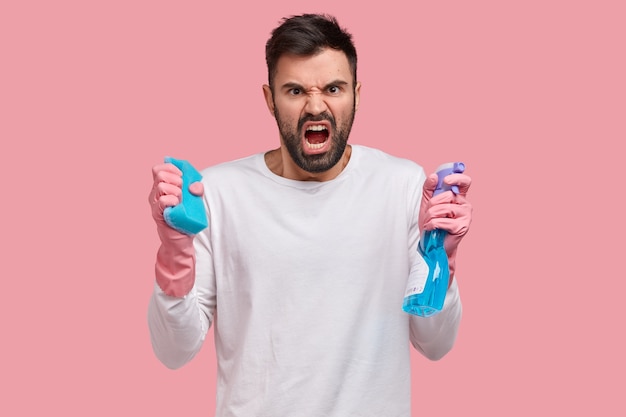 Studio shot van boze ongeschoren jongeman opent mond van woede, houdt spray en spons vast, fronst gezicht van ontevredenheid, drukt negatieve emoties uit