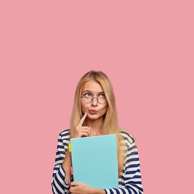 Studio shot van doordachte blonde vrouw portemonnees lippen, diep in gedachten, overdenkt