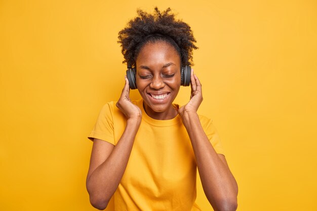Studio shot van gelukkig Afro-Amerikaanse tienermeisje houdt handen op koptelefoon geniet van perfecte geluidskwaliteit sluit ogen glimlacht breed gekleed in casual t-shirt geïsoleerd op levendig geel