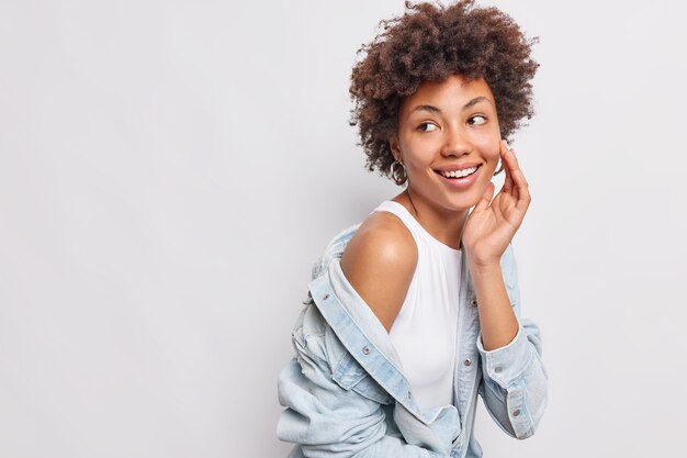 Studio shot van gelukkige vrouw houdt hand op gezicht kijkt terug met blije zorgeloze uitdrukking merkt iets aangenaams weg toont blote schouder draagt denim shirt met lange mouwen geïsoleerd op een witte muur