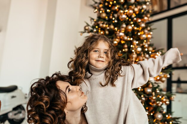 Studio shot van jonge mooie moeder en dochtertje met krullend haar dragen gebreide kleding poseren voor kerstboom