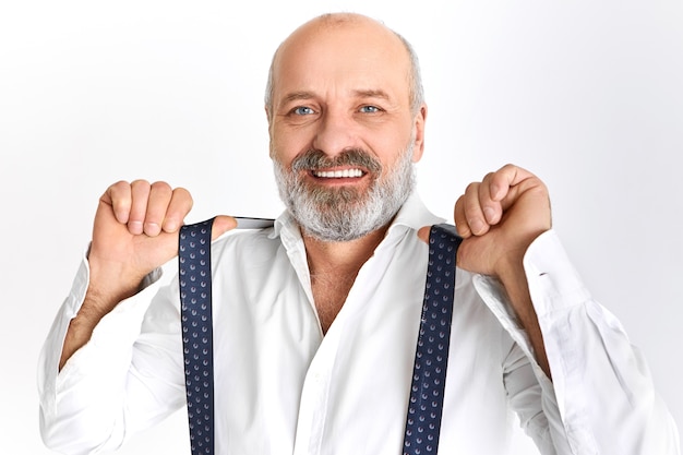 Studio shot van modieuze knappe bejaarde bebaarde man van in de zestig poseren geïsoleerd elegante kleding dragen, bretels aanpassen, glimlachend