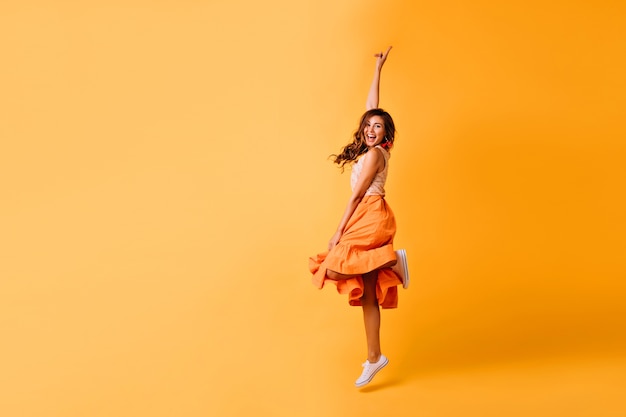 Studio shot van mooi meisje in oranje rok en witte schoenen. Opgewonden roodharige dame die op geel springt.
