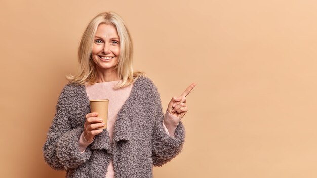 Studio shot van mooie blonde vijftig jaar oude vrouw glimlacht positief houdt wegwerp papieren kopje warme drank draagt bontjas geeft weg geïsoleerd over bruine muur