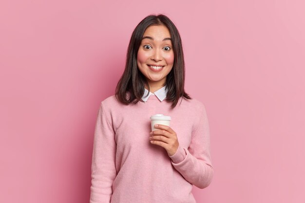 Studio shot van mooie brunette Aziatisch meisje heeft koffiepauze zachtjes glimlacht naar camera toont witte tanden gekleed in casual jumper poses in studio