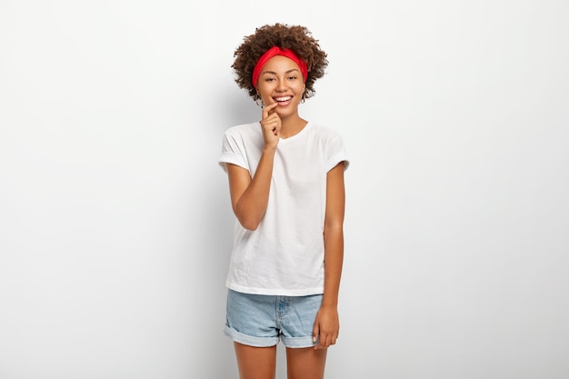 Studio shot van mooie gekrulde vrouw geniet van aangenaam moment, lacht zachtjes, draagt rode hoofdband, casual t-shirt en denim shorts, geïsoleerd op een witte achtergrond