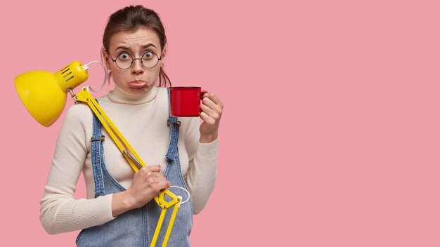 Studio shot van ontevreden jonge student portemonnees onderlip, koffie drinkt, bureaulamp gebruikt voor goed zicht