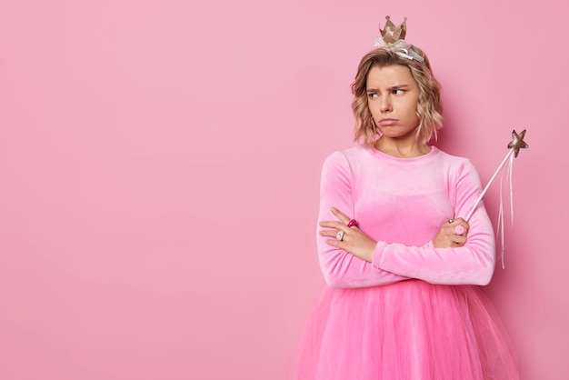 Studio shot van ontevreden jonge vrouw prinses draagt kleine kroon en jurk houdt toverstaf kijkt beledigd weg geïsoleerd over roze achtergrond lege ruimte voor uw advertentie of promotie.