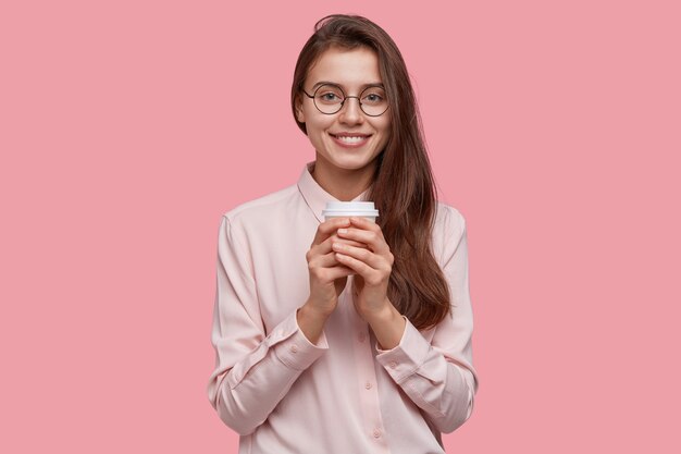 Studio shot van positieve schoolmeisje grijpt koffie om productief te werken, houdt papieren kopje drank, bezoekt café, draagt formeel overhemd