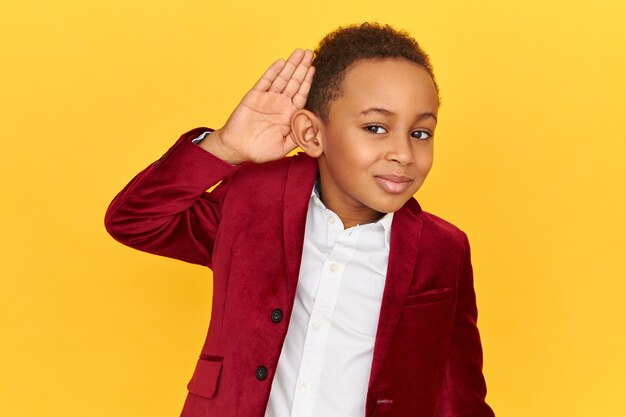 Studio shot van schattige snoopy donkere huid kleine jongen met een nieuwsgierige blik, met de handpalm tegen zijn oor om duidelijker te horen terwijl hij privégesprekken afluistert.