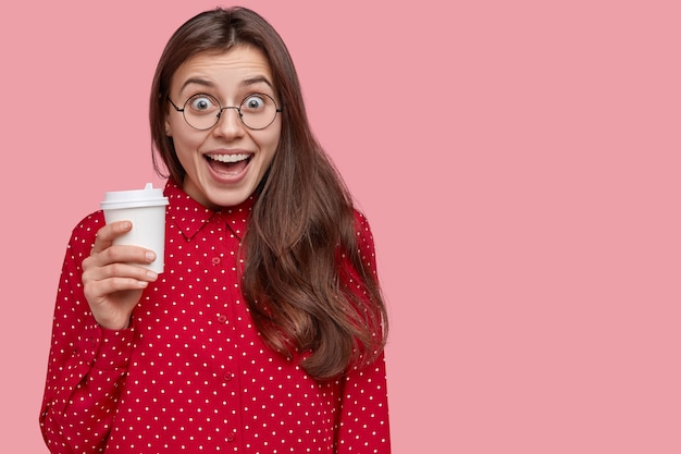 Studio shot van tevreden jonge vrouw lacht positief, drinkt afhaalkoffie, heeft pauze na lezingen